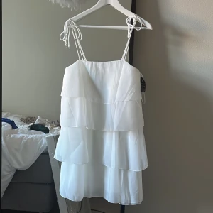 Vit volangklänning från Gina Tricot - Superfin vit klänning från Gina Tricot med flera lager volanger och smala axelband som knyts på axlarna. Perfekt för sommarens alla tillfällen och studenten. Lätt och luftig modell i syntetmaterial. Helt ny, aldrig använd.