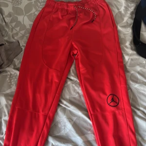 Röda Jordan mjukisbyxor med logga - Säljer ett par röda mjukisbyxor från Jordan med svart logga på benet. Byxorna har elastisk midja med dragsko och är tillverkade i ett mjukt syntetmaterial. Perfekta för en sportig och avslappnad stil. Inte använd och i jätte bra skick