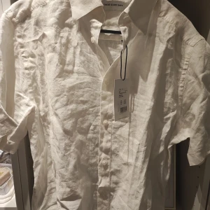 Vit linneskjorta These Glory Days Strl M - Snygg vit skjorta i linne från These Glory Days. Perfekt för varma dagar med sin lätta och luftiga känsla. Skjortan har korta ärmar och knappar framtill, vilket ger en klassisk look. Aldrig använd, slimfot storlek M