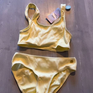 NY! Gul bikini från KappAhl strl 146/152 - HELT NY  och oanvänd gul bikini från KappAhl med ribbad textur. Toppen har breda axelband och en sportig look. Strl 146/152. Nypris: 298 kr, nu 95kr!