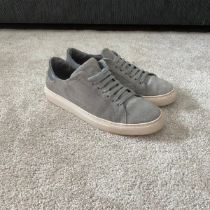 Axel Arigato Clean 90 Grå Suede Sneakers - Snygga grå sneakers från Axel Arigato i mocka med snörning. Perfekta för en stilren look. De har en rund tå och en bekväm sula som ger bra stöd. Passar utmärkt för både vardag och avslappnade tillfällen. Sulorna har bytts ut till ett par från ecco och orginalsulorna har tappats bort annars i fint skick.