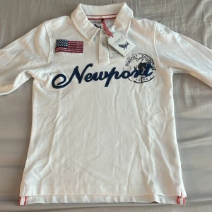 Vit pikétröja från Newport - Vit pikétröja från Newport med broderad amerikansk flagga på bröstet och stor blå logga framtill. Klassisk krage med knappar och röda detaljer på insidan. Perfekt för en sportig och stilren look.