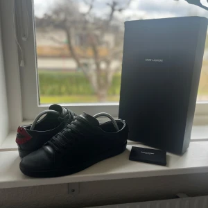 Saint Laurent skor - Svarta sneakers från Saint Laurent med snörning och rund tå. Baksidan har en unik röd och svart läppdetalj. Väldigt bra skick och extremt snygga nu inför våren/sommarn
