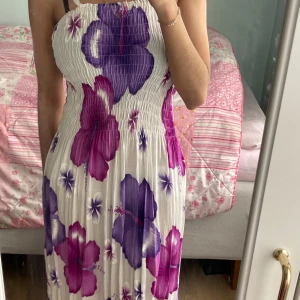 Vit långklänning med lila och rosa blommor - Säljer en somrig långklänning med stora lila och rosa blommor på vit botten. Klänningen har smock upptill och är ärmlös med axelband. Perfekt för varma dagar och midsommarfirande.