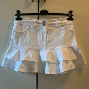 Vit kjol med volang från Zoul - Säljer en söt jeans vit kjol från Zoul med volangdetaljer. Kjolen har en knapp och dragkedja framtill samt fickor. Perfekt för sommardagar! 🌸priset kan diskuteras🫶🏼