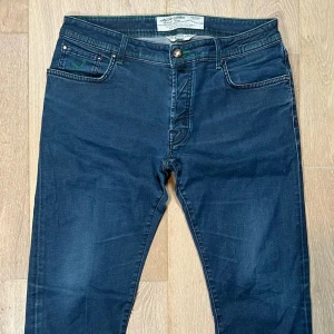 Blå slim fit jeans från Jacob Cohen - Säljer ett par blå jeans från Jacob Cohen med snygga gröna sömmar och broderad J på fickan. Klassisk femficksmodell med knappgylf och patch med J på baksidan. Perfekta för dig som gillar detaljer och stilren design. Pris kan diskuteras vid snabb affär!