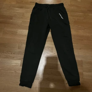 Svarta mjukisbyxor med tryck från Björn Borg - Svarta mjukisbyxor från Björn Borg med vitt tryck på ena fickan. Byxorna har resår och snörning i midjan samt muddar vid bensluten. Perfekta för en avslappnad stil.