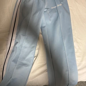 Ljusblå byxor från Palm Angels - Snygga ljusblå byxor från Palm Angels med vita och svarta ränder längs sidorna. Byxorna har en avslappnad passform och är perfekta för en trendig look. Märkesloggan är tryckt på framsidan.