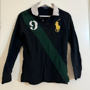 Svart pikétröja från Polo Ralph Lauren med grön detalj - Säljer en svart långärmad pikétröja från Polo Ralph Lauren med vit krage, grön diagonal rand och stor gul broderad logga på bröstet. Siffra 9 på axeln. Klassisk och stilren modell, perfekt till många tillfällen.