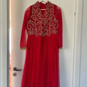 Röd långklänning med broderier - Säljer en röd långklänning med v-ringning och vackra broderade detaljer i guld och rosa på överdelen. Klänningen har transparenta långa ärmar med guldkant och medföljande sjal och byxor som är justerbara. Perfekt för bal eller fest. Den ger indisk vibes.
