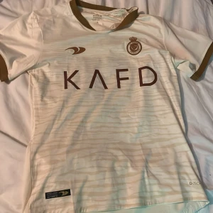 Al Nassr Ronaldo 7 fotbollströja - Vit kortärmad Al Nassr fotbollströja med bruna detaljer och tryck. Stort klubbmärke på bröstet, sponsorlogga och Ronaldo 7 på ryggen. Tillverkad i lätt och ventilerande material, perfekt för match eller träning.