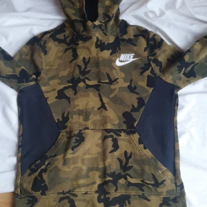 Nike hoodie med camouflage-mönster - Säljer en Nike hoodie i camouflage med broderad vit logga på bröstet. Tröjan har huva, lång ärm och känguruficka framtill. Materialet är mjukt och bekvämt, perfekt för en avslappnad stil.