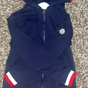 Mörkblå hoodie från Moncler med dragkedja - Säljer en mörkblå hoodie från Moncler med dragkedja och huva. Jackan har vita och röda detaljer på huvan och ärmslut samt Moncler-logga på ärmen. Perfekt för en sportig och stilren look.
