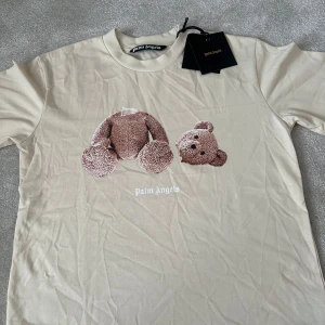 Beige t-shirt med nalletryck från Palm Angels - Säljer en beige t-shirt från Palm Angels med ikoniskt nallebjörn-tryck på framsidan och stor vit logga på ryggen. T-shirten har rund hals och är kortärmad. Perfekt statement-plagg för dig som gillar streetwear. Storlek S men passar även M