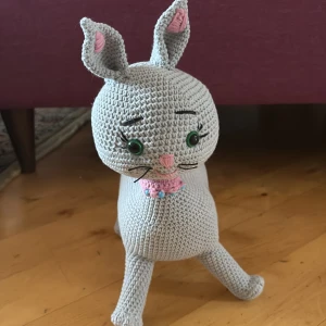 Virkad katt i grått garn - Handgjord virkad katt i ljusgrått garn med gröna plastögon, rosa nos och detaljer, samt en rosa virkad rosett runt halsen. Katten har långa ben, spetsiga öron och en lång svans. Perfekt som dekoration eller gosedjur.