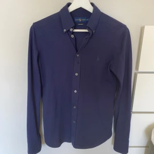 Mörkblå skjorta från Ralph Lauren - Snygg mörkblå långärmad skjorta från Ralph Lauren i modellen Knit Oxford. Skjortan har knappar framtill och en broderad logotyp på bröstet. Perfekt för en stilren look. Är i nyskick.Hör av er vid funderingar