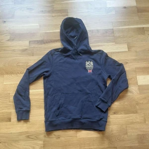 Morris hoodie  - Säljer en hoodie från Morris med broderad logga och brittisk flagga på bröstet. Tröjan har huva med snörning, känguruficka och ribbade muddar. Perfekt för dig som gillar klassisk stil med en sportig touch.
