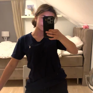 Mörkblå t-shirt från Ralph Lauren - Mörkblå t-shirt från Ralph Lauren med klassisk v-ringning och vit broderad logga på bröstet. Skön passform och tillverkad i mjuk bomull. Perfekt basic-plagg med stilren look och korta ärmar. Använd fåtal gånger!