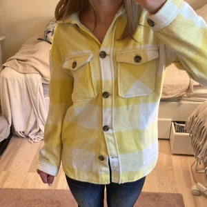 Gul och vit rutig overshirt från H&M - Kappa/overshirt från H&M i storlek 158, med ett gul och vit rutigt mönster. Jackan har två bröstfickor med knappar, klassisk krage, knapp vid ärmens slut och knäppning framtill. Använd men bra skick.