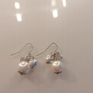 Söta örhängen med pärlor och hjärtan - Charmiga örhängen med stora vita pärlor, små silverfärgade hjärtan och gnistrande stenar. Hängande modell med metallkrokar och fina detaljer i botten av pärlorna. Perfekta för dig som gillar en söt och romantisk stil.