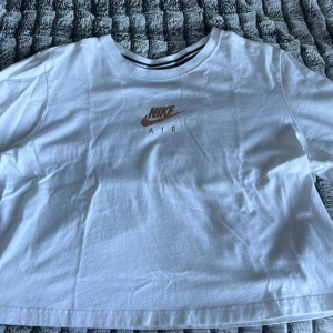 Vit Nike Air t-shirt i bomull - Vit t-shirt från Nike Air med klassisk logga i guldigt framtill och text på ryggen. Modellen är kortärmad och har en avslappnad passform. Tillverkad i mjuk bomull, perfekt för en sportig och clean look.