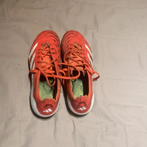 Adidas Predator röda fotbollsskor - Säljer ett par Adidas Predator fotbollsskor i rött med vita detaljer och svarta loggor. Skorna har snörning, klassisk låg profil och mönstrad ovandel för extra grepp. Perfekta för dig som vill sticka ut på planen med stilrena och sportiga skor.