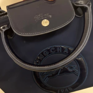 Mörkblå handväska från Longchamp - Snygg mörkblå handväska från Longchamp med glansiga skinnhandtag och lock. Väskan har ett stort präglat Longchamp-logotyp framtill och stängs med dragkedja och tryckknapp. Perfekt storlek för att bära med sig det viktigaste.