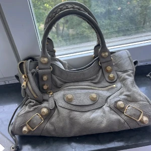 Beige Balenciaga City handväska i skinn - Säljer en beige Balenciaga City handväska i mjukt skinn med guldiga detaljer och dragkedjor. Väskan har flätade handtag, dekorativa nitar och spännen samt en spegel. Perfekt för dig som vill ha en ikonisk och rymlig väska med edgy vibe.