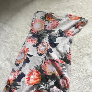 Grå blommig kortklänning - Superfin grå kortklänning med stora färgglada blommor i rosa, orange och grönt. Klänningen har rund halsringning och korta ärmar. Perfekt för dig som gillar ett statement-plagg med mycket färg och mönster.