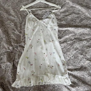 Vit blommig sommarklänning H&M - Supersöt vit kortklänning från H&M med tunna axelband och volangkant nertill. Klänningen har ett diskret blommigt mönster i pastellfärger och en lätt, somrig känsla. Perfekt för varma dagar och har en feminin, romantisk stil.