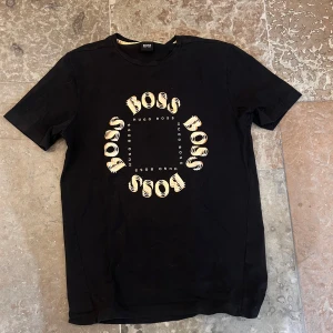 Svart BOSS t-shirt med tryck - Svart t-shirt från Hugo Boss med coolt cirkelformat BOSS-tryck i gult och vitt på bröstet. Klassisk rund halsringning och korta ärmar. Skön bomullskvalitet som passar perfekt till jeans eller shorts.