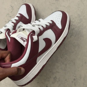 Två Nike Dunk Low  - Två par Nike Dunk Low sneakers. Ena paret är i vinrött och vitt skinn med klassisk swoosh och vita snören. Andra paret har denim-look i blå och grå toner med vit swoosh och blå snören, samt retrovit sula. Båda har rund tå och platt sula – perfekta för dig som gillar streetstyle. Säljer båda för 700kr men seperat 400kr  Båda är storlek 42,5   Kan mötas upp vart som stockholm