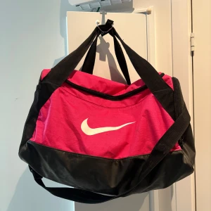 Rosa och svart Nike träningsväska - Säljer en sportig träningsväska från Nike i stark rosa med svarta detaljer och stort vitt logotryck. Väskan har flera fack, dragkedja och breda axelband. Perfekt storlek för gymmet eller sportaktiviteter. Materialet är slitstark polyester och syntet.