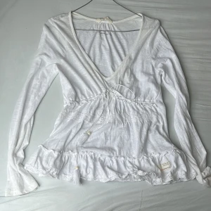Vit blus med volang från Odd Molly - Supersöt vit blus från Odd Molly med v-ringning, långa ärmar och knyt i midjan. Blusen har volangkant nertill och spetsdetaljer. Tillverkad i mjuk bomull och perfekt för dig som gillar en avslappnad stil. Man kan  knyta snöret där fram så att blusen sitter tajtare runt överdelen. Den lite gul vid v-ringningen, men själv skulle jag säga att det inte märks av. Priset kan diskuteras❤️