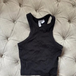 Svart croppad linnetopp H&M Divided - Svart ribbad croppad linnetopp från H&M Divided. Modellen har breda axelband och hög halsringning, perfekt att styla med jeans eller kjol. Toppens material är mjuk bomull med stretch för en bekväm passform.