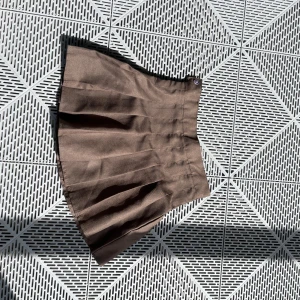 Brun plisserad kortkjol - Snygg brun plisserad kortkjol med klassisk siluett. Kjolen har tydliga veck och är tillverkad i ett slätt, lite glansigt tyg som ger en stilren look. Perfekt att matcha med en oversized hoodie eller en tight topp för en trendig vibe.