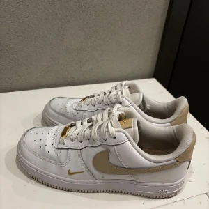 Nike Air Force 1 vit/guld sneakers - Nike Air Force 1 sneakers i vitt skinn med snygga gulddetaljer på swoosh, häl och snörning. Klassisk låg modell med perforerad tå och platt sula. 