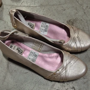Ballerinaskor - Snygga beige ballerinaskor med kilklack och rund tå. Skorna har dekorativa remmar framtill med små guldfärgade nitar och en mjuk rosa innersula. Perfekta för dig som vill ha en klassisk men ändå trendig look.