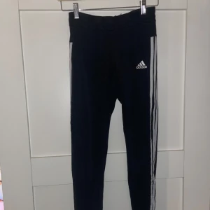 Svarta Adidas tights  - Snygga svarta tights från Adidas med klassiska vita ränder längs sidorna och Adidas-logga på låret. Perfekta för träning eller chill, med tight passform och stretchigt material som ger skön rörelsefrihet.
