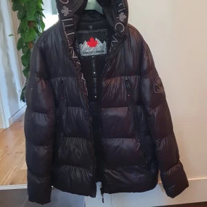 Svart dunjacka från Zavetti Canada - Snygg svart dunjacka från Zavetti Canada med stor huva och feta loggor längs kanten. Jackan är quiltad och har dragkedja framtill samt flera fickor med dragkedja. Perfekt för kalla dagar när du vill hålla stilen och värmen. Mindre skada på höger ärm. Storlek M.
