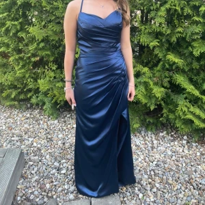 Mörkblå satin balklänning  - Säljer denna jättefina balklänning som jag själv faktiskt använde. Den är då använd 1 gång och är fortfarande i väldigt bra skick. Perfekt alternativ för balen men kan även användas som en klänning för andra finare tillfällen. På ryggen har den även en fin knytning som ger hela klänningen en extra touch. Botten har syts upp för att passa mig som är 167+ klackar. Har även del för att kunna hålla upp klänningen längst ner, som synligt på bilden. Pris kan i ddtta tillfälle diskuteras.