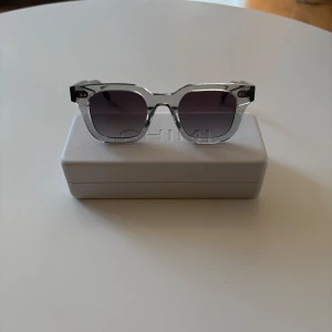 Grå transparenta solglasögon CHIMI - Snygga solglasögon från CHIMI med transparent grå båge och fyrkantig form. Glasen är mörktonade och ger en cool vibe. Kommer med originaletui i vitt. Perfekta för att lyfta din stil under soliga dagar.