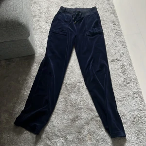 Mörkblå velourbyxor Juicy Couture XS - Mörkblå mjukisbyxor från Juicy Couture i glansig velour. Byxorna har snörning i midjan, fickor fram och bak med knappar samt bred resår i midjan. Perfekta för en chill och trendig look.