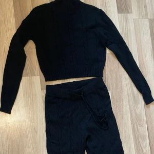 Svarta stickade sett från Kaylla - Svarta stickade byxor och tröja från Kaylla med bred resår och snörning i midjan. Byxorna har ett kabelstickat mönster och är i mjukt material med loose passform och raka ben. Perfekta för en chill och avslappnad look.