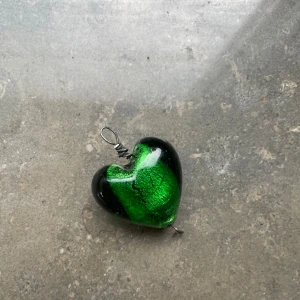 Grönt hjärthänge i glas - Unikt hjärthänge i glas med intensivt grön och svart färg. Hängsmycket har en cool metallögla upptill för att enkelt fästa på en kedja eller band. Perfekt för dig som vill sticka ut med ett personligt smycke.