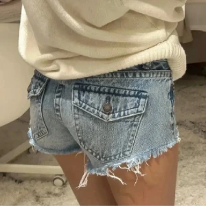 Blå jeansshorts från Pull&Bear - Snygga blå jeansshorts från Pull&Bear med råa, fransiga kanter och slitna detaljer. Klassisk femficksmodell med knappar och bälteshällor. Perfekta för varma dagar när du vill ha en avslappnad och trendig look.