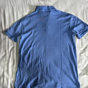 Blå pikétröja Ralph Lauren slim fit - Snygg ljusblå pikétröja från Ralph Lauren i modellen Custom Slim Fit. Tröjan har klassisk krage, tre knappar och den ikoniska broderade polospelaren på bröstet. Tillverkad i mjuk bomull och har korta ärmar. Perfekt för en clean och stilren look. Helt oanvänd. Pris kan diskuteras 