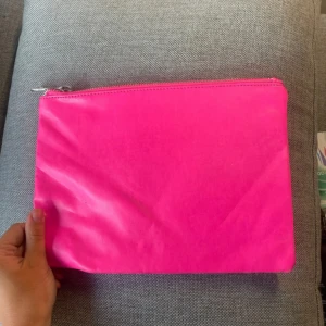 Neonrosa kuvertväska med dragkedja - Enkel och snygg kuvertväska i neonrosa färg med dragkedja upptill. Perfekt att ha med sig när du vill sticka ut lite extra. Väskan är slät och har en stilren design utan mönster eller logga.