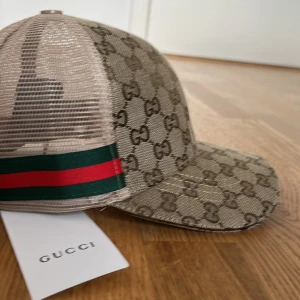 GG keps  - Snygg keps med klassiskt GG-monogram i beige och brunt. Kepsen har mesh på baksidan och en grön-röd randig detalj på sidan. Perfekt accessoar för att lyfta din outfit med lyxig känsla.