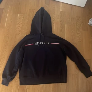 Svart hoodie Tommy Hilfiger M - Mörk blå hoodie från Tommy Hilfiger i storlek M. Klassisk design med broderad Hilfiger-logga och röd-vita detaljer på bröstet. Tröjan har huva med snörning och långa ärmar. Perfekt för en chill och stilren look.